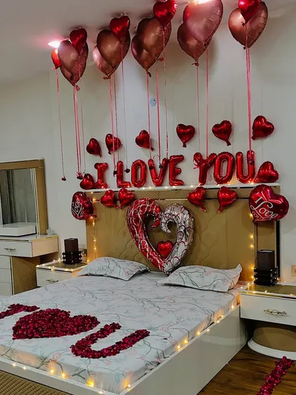 A close-up of the bed decoration, featuring "I LOVE YOU" spelled out with rose petals, heart-shaped foil balloons, and a canopy of red chrome balloons. The warm fairy lights around the headboard add a soft, romantic glow.