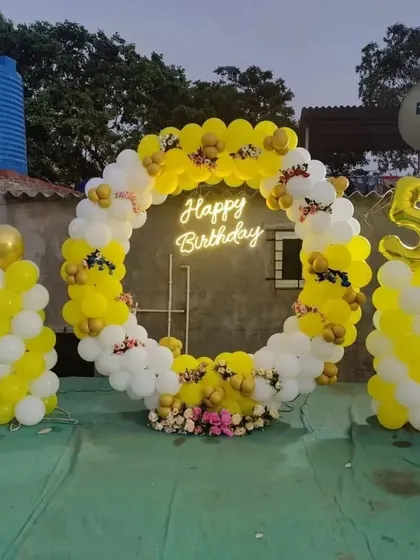 A birthday party on a terrace with a circular balloon arch in yellow and white, and a 'Happy Birthday' neon sign.