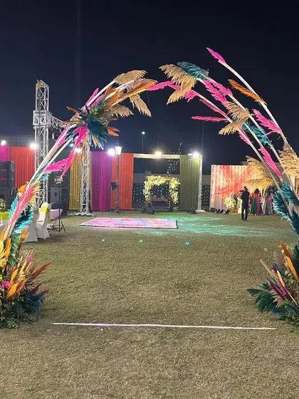 A wide view of an outdoor Mehendi setup. The lawn is transformed with a colourful entrance arch, vibrant backdrops for seating areas, and a spacious dance floor for an evening of celebration.