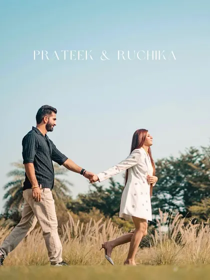 A stylish shot of the couple walking through a field of tall grass under a clear blue sky. This pre-wedding photo has a chic, editorial feel.