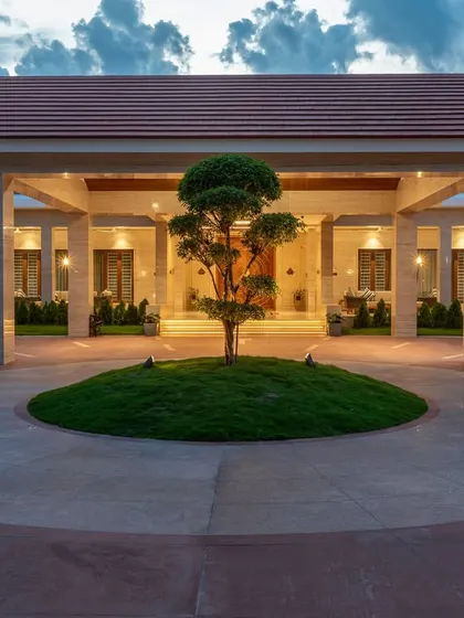 The main portico at dusk, illuminated to create a grand and welcoming entrance. The design uses classical pillars and a circular driveway centered around a bonsai tree, establishing a formal yet serene sense of arrival.