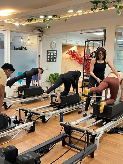 An instructor provides hands-on assistance during a group reformer class, ensuring proper alignment for a leg-back exercise on the box.