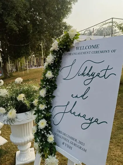 A chic and modern welcome sign for an engagement ceremony. The elegant calligraphy with the couple's names and event details is beautifully accented with a garland of fresh white flowers.