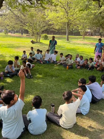 Children sit in a circle in a park for an outdoor lesson, learning about their environment in a natural, engaging setting.