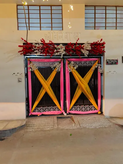 A close-up of the decorated entrance gate of the wedding house. The doors are adorned with bright pink and yellow drapes and floral arrangements.