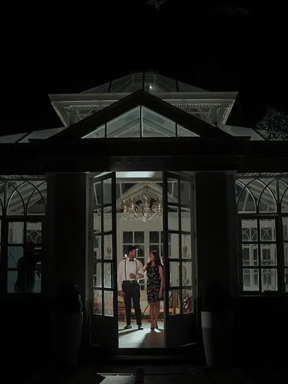 A striking silhouette of a couple sharing an intimate moment in the doorway of a grand, lit-up glasshouse at night. This shot uses architectural framing to create a sense of romance and privacy.