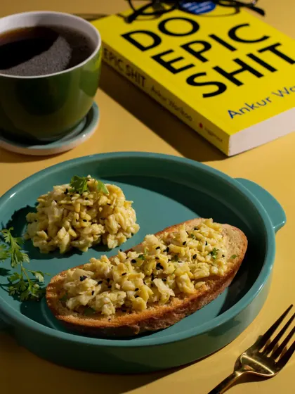 A simple, classic breakfast of scrambled eggs on toast. The bright, cheerful styling with a book and a cup of black coffee creates a relatable and appealing morning scene.
