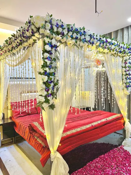 A beautiful wedding night setup with a floral arch of white and blue flowers. The bed is draped with white curtains and lit by fairy lights.
