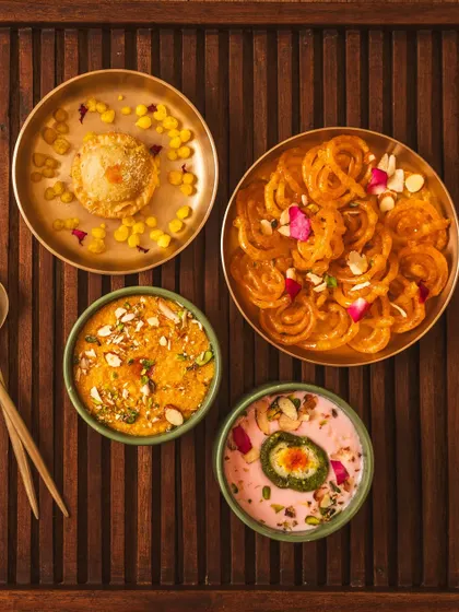 A flat lay of traditional Indian desserts like jalebi and ghevar, styled with brass plates and a dark wood background. This moody, festive shot evokes a sense of celebration and rich flavor.