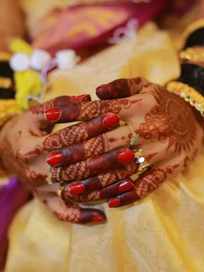 A close-up of a bride's hands, showing her red nail polish, henna design, and traditional bangles.