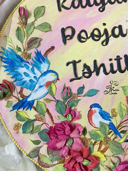 A close-up of the "Kalyan, Pooja, Ishitha" nameplate, showing the detailed blue bird with spread wings among the sculpted roses.