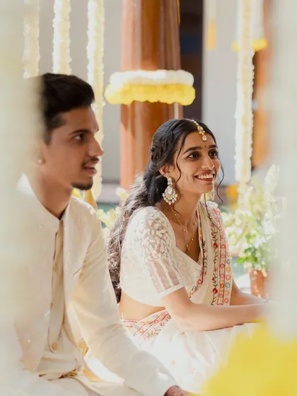 The couple shares a sweet moment during their Haldi ceremony, dressed in elegant white outfits that provide a beautiful contrast to the yellow turmeric.