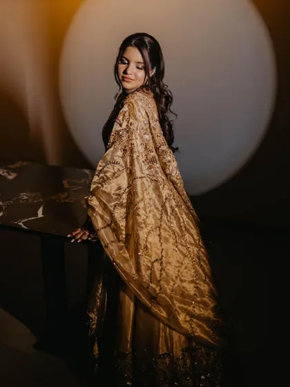 A stunning portrait of the bride in her golden Sangeet outfit. The lighting creates a halo effect, making her look ethereal and radiant.