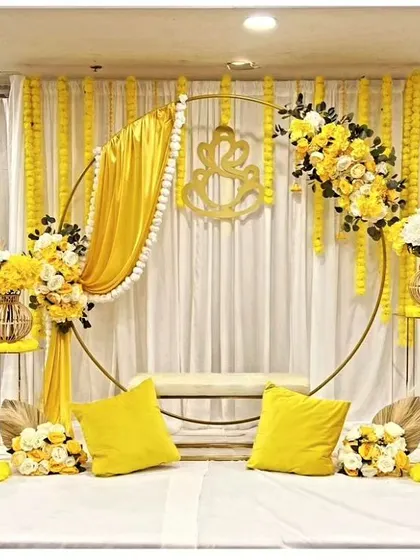 A modern and chic Haldi setup featuring a golden ring backdrop draped with yellow fabric and adorned with floral arrangements. The auspicious Ganesha motif adds a traditional touch to the contemporary design.