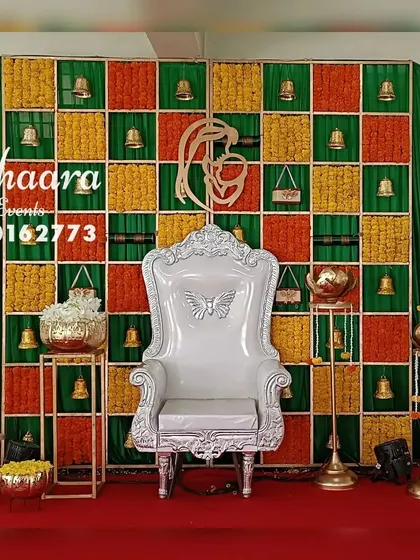 A traditional baby shower stage with a unique backdrop. The grid features squares of marigolds and hanging bells, with a mother-and-child motif, blending classic elements with a modern design.