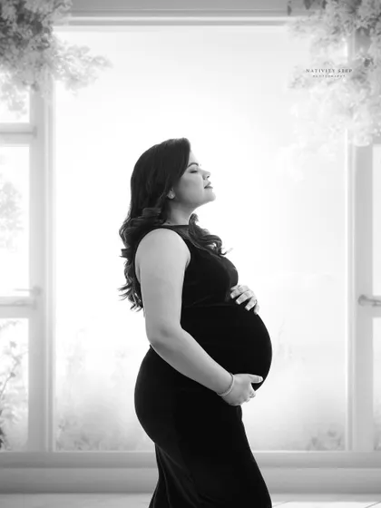 A serene black and white portrait by a window. The soft, natural light and the mother's peaceful expression create a feeling of calm and anticipation.