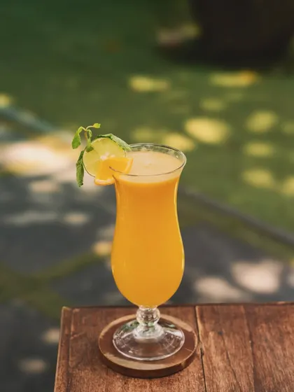 A classic mango lassi or smoothie in a shapely glass, set on a wooden coaster outdoors. The soft, natural light and blurred green background create a serene and inviting mood.