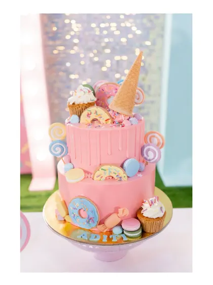 A spectacular two-tiered cake for a candyland party. It's dripping with pink icing and adorned with donuts, lollipops, macarons, and an upside-down ice cream cone.
