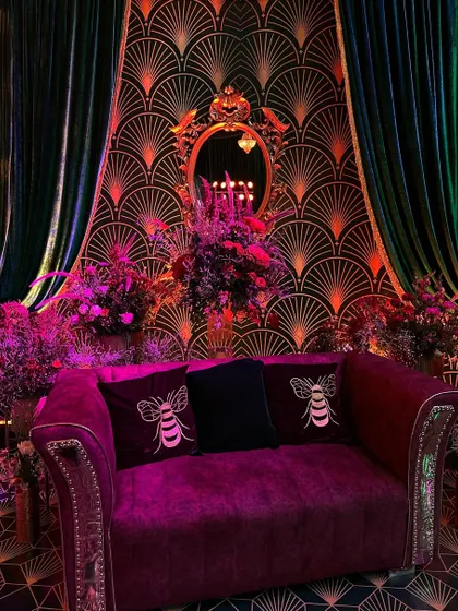 A sophisticated lounge area with a vintage, art-deco feel. A plush magenta sofa sits before a patterned wall, framed by emerald velvet curtains and an ornate golden mirror.