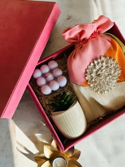 A chic and aesthetic Diwali hamper in a luxe pink box. It contains a bubble candle, a small succulent in a ceramic pot, and a traditional Gota potli bag filled with trail mix.