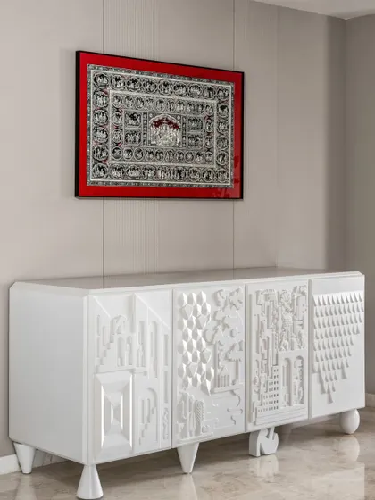 This custom-designed sideboard is a piece of functional art. The white lacquered surface features intricate 3D patterns on each door, creating a fascinating play of light and shadow. Above it, a traditional Indian Pichwai artwork in a bold red frame provides a stunning cultural contrast.