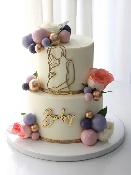 An elegant two-tiered baby shower cake with a "mom-to-be" silhouette, fresh flowers, and a cascade of colorful chocolate spheres.