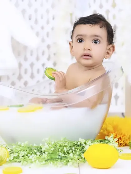 A baby sits in a milk bath, holding a slice of lime and looking at the camera. The bright colors of the citrus pop against the white milk.