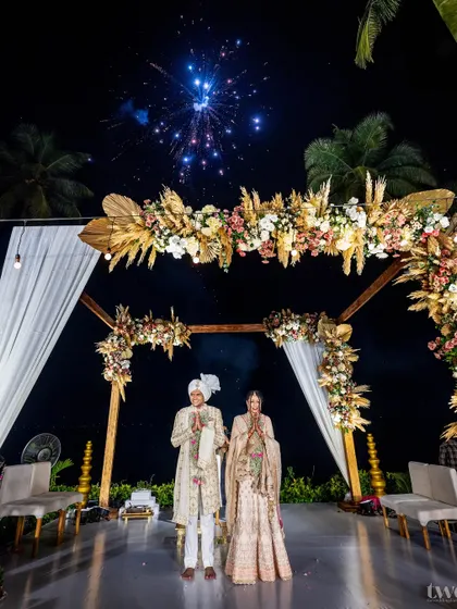 The couple stands under their wedding mandap against a backdrop of fireworks. A spectacular and celebratory portrait from their destination wedding in Goa.
