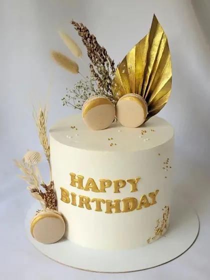 An elegant design in ivory and gold. This cake is decorated with gold lettering, macarons, and a beautiful arrangement of dried wheat and palm leaves for a rustic-chic look.