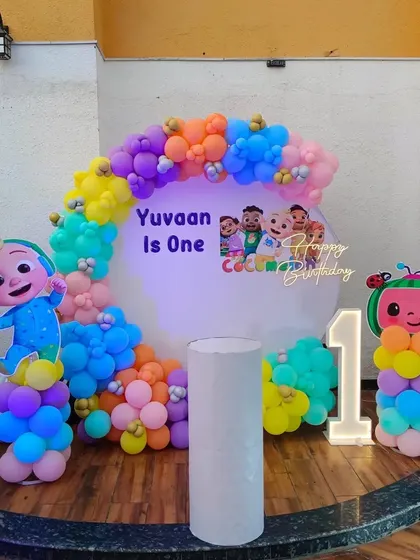 A clean and bright Cocomelon first birthday decoration with a white circular backdrop, light-up number, and colorful balloon arch.