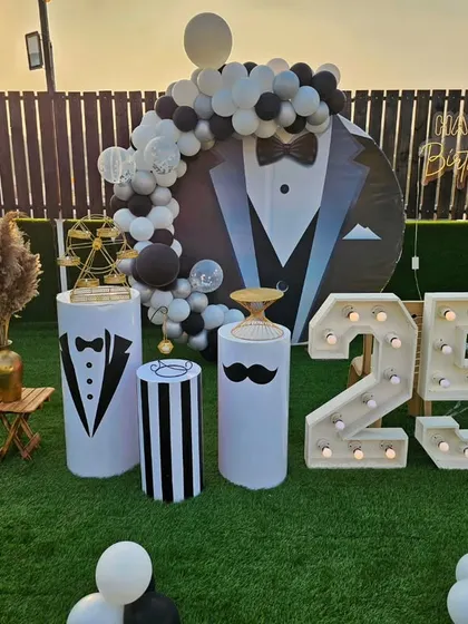 A dapper "tuxedo" themed 25th birthday party. This outdoor setup features a suit-and-tie backdrop, marquee numbers, and fun details like a mustache prop.
