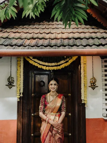 This classic South Indian bridal portrait captures the bride's elegance in front of a beautifully decorated traditional doorway, highlighting her stunning attire and poise.