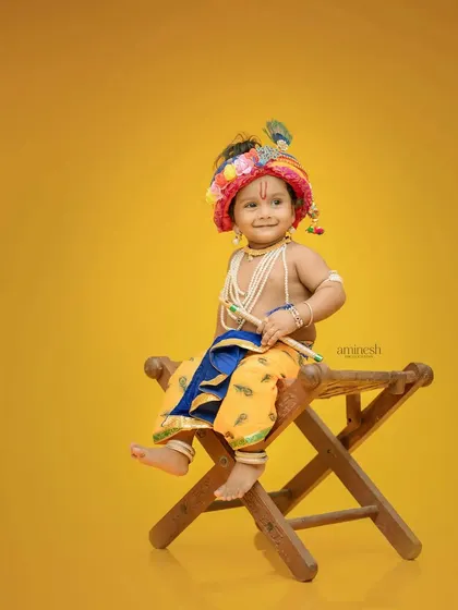 A simple and classic portrait of a baby dressed as Krishna, seated on a wooden stool. The solid yellow background makes the traditional costume pop.