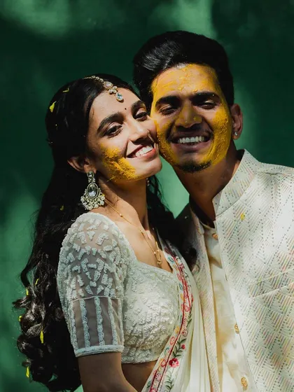 A heartwarming portrait of the couple, their faces glowing with happiness and turmeric paste, against a rich green background.