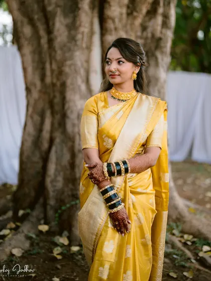 A poised and elegant portrait of the bride in her yellow saree, standing by a large tree.