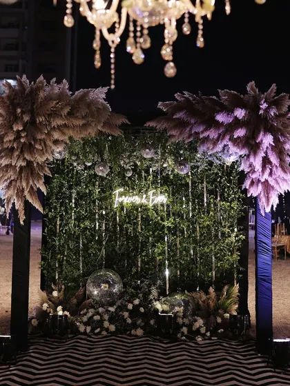 A stylish photo booth with a "Forever & Ever" neon sign on a green wall, framed by pampas grass and disco balls.