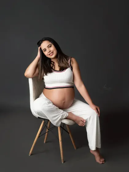 A relaxed and chic studio portrait. This mom-to-be is sitting comfortably in a modern chair, wearing a stylish white two-piece outfit that highlights her bump.