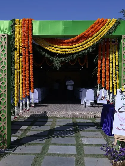 An outdoor entrance decoration for a ceremony, featuring a green arch with hanging marigold garlands. A custom welcome sign greets the guests, setting a festive tone.