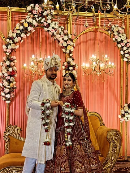 This is a final wedding stage setup during the Jaimala ceremony. The decor includes golden arches with white and pink floral arrangements, warm chandelier lighting, and a comfortable sofa for the couple.