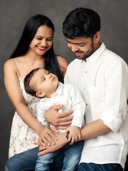 A classic family portrait against a simple, dark backdrop. The focus is entirely on the connection and love between the parents and their adorable little one.
