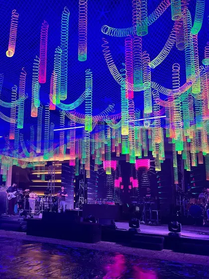 A wide view of the main stage and ceiling installation for a neon-themed corporate party. The hanging neon slinkies covered the entire ceiling, creating a colorful canopy over the band and dance floor.