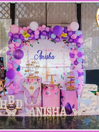 A full-stage butterfly-themed fifth birthday. The purple and pink decor features a 'Our Little Anisha' backdrop, large butterfly cutouts, and a 'ANISHA' marquee.