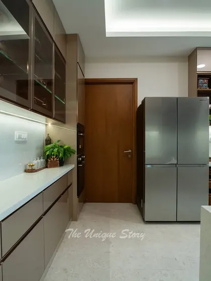 A parallel kitchen layout with ample counter space and storage. The design includes glass-front upper cabinets to display crockery and a large side-by-side refrigerator, all illuminated by soft LED strip lights.