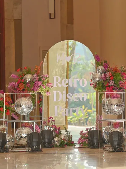 The entrance to the "Retro Disco Party" Mehendi, featuring a mirrored arch with the event's theme, surrounded by disco balls and lush, colorful flowers.