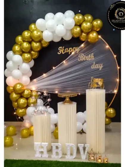 This design shows how we mix textures for a unique look. A semi-arch of gold and white balloons is paired with sheer, fairy-lit fabric across a gold ring frame, creating a soft yet celebratory backdrop for a birthday.