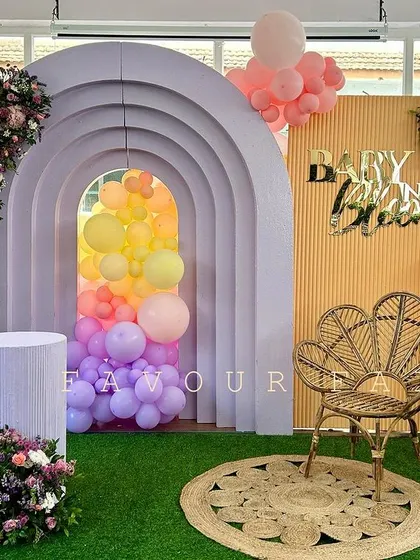 A 'Baby in Bloom' baby shower with a rainbow of pastel colors. The setup features a unique arched backdrop with a balloon-filled tunnel effect, a wicker peacock chair, and beautiful floral arrangements.
