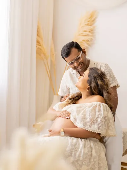 A tender moment captured in our natural light studio. The couple shares a laugh, with the mother-to-be seated and looking up at her partner, her lace dress and the soft background creating a dreamy, ethereal vibe.