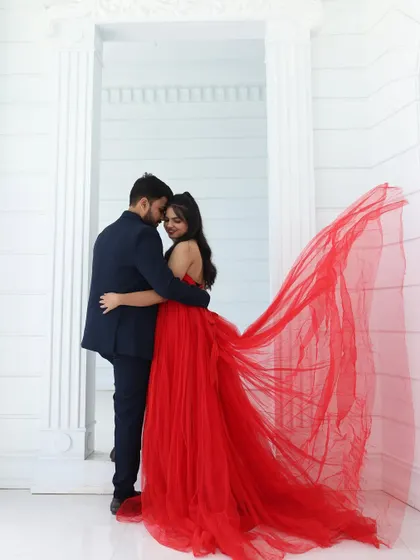 A romantic pre-wedding embrace in a studio with an all-white background. The flowing red trail of the gown provides a stunning pop of color.