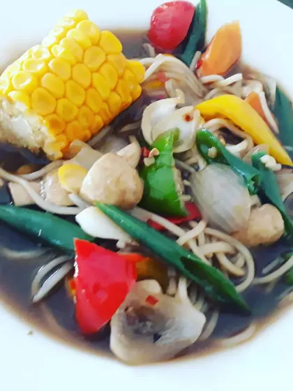 A comforting Chinese-style soupy noodle bowl, loaded with fresh vegetables like bell peppers, mushrooms, and a whole corn on the cob.