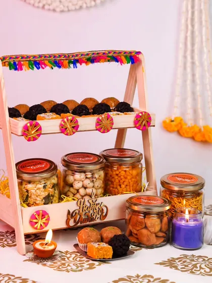 A vibrant and playful Diwali hamper featuring a two-tiered wooden stand. The stand is filled with jars of namkeen, sweets, and other snacks, decorated with colorful tassels. It's a gift that doubles as a beautiful centerpiece.